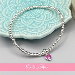 May include: A sterling silver bracelet featuring small, round silver beads and a pink heart-shaped charm. The bracelet is displayed on a light blue decorative plate. The text "Sterling Silver" is displayed in a pink banner.