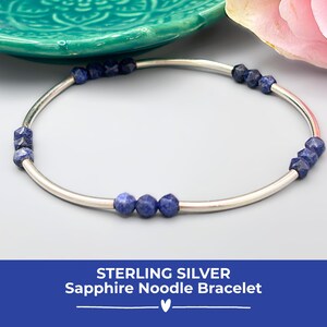 May include: A sterling silver sapphire noodle bracelet. The bracelet has a silver tube design with dark blue sapphire beads. The background includes a teal plate and a pink flower. The text on the image reads: Sterling Silver Sapphire Noodle Bracelet.