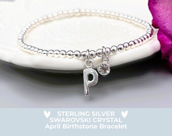 May include: A silver beaded bracelet with a dangling letter "P" charm and a small crystal charm. The bracelet is displayed on a white plate with a blurred purple flower in the background. Text at the bottom reads "STERLING SILVER SWAROVSKI CRYSTAL April Birthstone Bracelet."