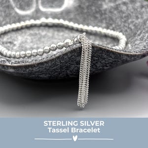 May include: A silver bracelet with a beaded design and a tassel detail. The bracelet is presented in a grey, textured bowl. The text "STERLING SILVER Tassel Bracelet" is at the bottom.