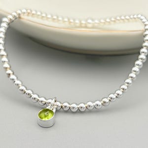 May include: A stretch bracelet composed of small, polished silver-toned spherical beads arranged in a continuous circular loop. A single, small, round, light green gemstone charm is suspended from the bracelet by a silver-toned metal bail.
