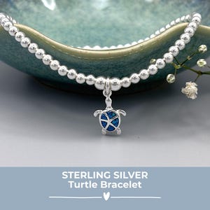 May include: A sterling silver turtle bracelet. The bracelet is made of small, round silver beads. A turtle charm with a blue and silver design hangs from the centre. The image is set against a soft blue background.