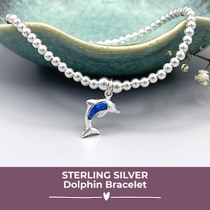 May include: A sterling silver bracelet featuring small silver beads and a dolphin charm with a blue inlay. The bracelet is presented in a teal bowl. The text at the bottom reads "STERLING SILVER Dolphin Bracelet."