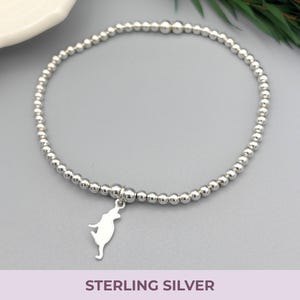 May include: A stretch bracelet composed of small, polished sterling silver beads arranged in a circular band. A flat, silver-toned charm shaped like a cat in a leaping or playful pose hangs from the center of the bead strand.