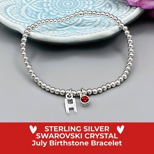 May include: A silver beaded bracelet with a letter "H" charm and a red Swarovski crystal charm. The bracelet is presented on a light blue plate. The text at the bottom reads "STERLING SILVER SWAROVSKI CRYSTAL July Birthstone Bracelet."
