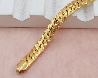May include: A gold chain bracelet with a textured, link-style design. The bracelet is lying on a white and pink surface.