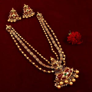 Peut inclure: Un collier en or avec un pendentif rouge, vert et blanc. Le collier a trois rangs de cha&icirc;ne en or avec de petites perles rondes. Le pendentif est en forme d'&eacute;ventail et pr&eacute;sente des d&eacute;tails complexes. Le collier est sur un fond rouge.