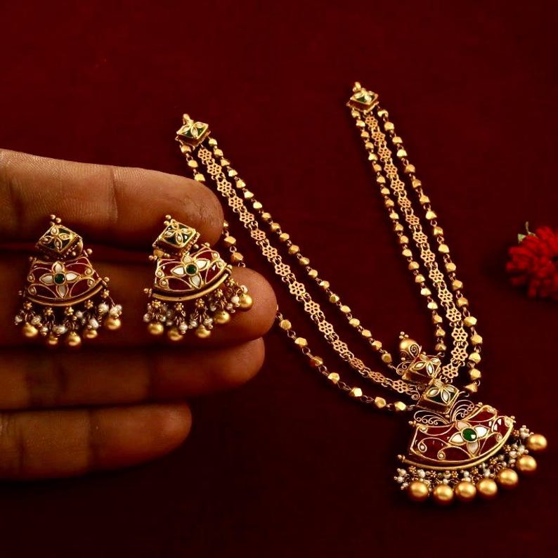 Peut inclure: Un collier en or avec trois rangs de cha&icirc;ne et un pendentif en &eacute;mail rouge, vert et blanc. Le pendentif est en forme de demi-lune et pr&eacute;sente un motif floral. Le collier est accompagn&eacute; de boucles d'oreilles assorties.