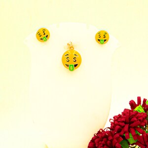 May include: A gold-tone necklace and earrings set featuring a yellow smiley face pendant with a green dollar bill sticking out of its mouth. The pendant and earrings have black eyes and a black outline.