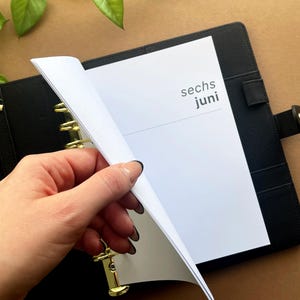 Könnte beinhalten: Schwarzer Lederplaner mit goldfarbenen Ringen und Akzenten. Der Planer ist geöffnet und zeigt eine weiße Seite mit dem Text "sechs juni" in dunkelgrauer Schrift. Eine Hand blättert um.