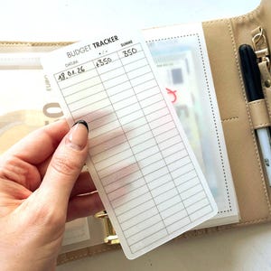 Könnte beinhalten: Ein beiger Budgetplaner mit einer transparenten Plastiktasche, die Bargeld enthält. Ein weißes Budget-Tracker-Blatt wird davor gehalten, mit den Worten "BUDGET TRACKER" und Spalten zur Verfolgung von Einnahmen und Ausgaben. Ein schwarzer Stift befindet sich in einem Halter.