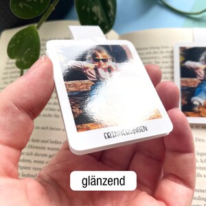 Ein personalisierbares magnetisches Lesezeichen mit Foto eines Kleinkinds wird so in die Kamera gehalten, dass die glänzende Oberfläche gut zu erkennen ist. Darunter steht in einem weißen Feld der Text „glänzend".
