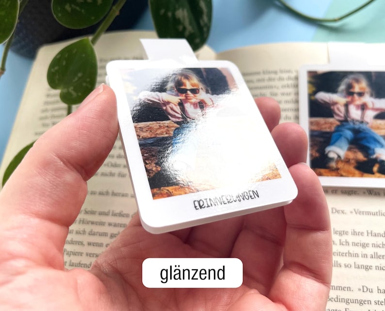 Ein personalisierbares magnetisches Lesezeichen mit Foto eines Kleinkinds wird so in die Kamera gehalten, dass die glänzende Oberfläche gut zu erkennen ist. Darunter steht in einem weißen Feld der Text „glänzend".