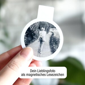 Ein magnetisches Lesezeichen mit Foto einer Person, die von hinten zu sehen ist, wird vor einem unscharfen Hintergrund in einer Hand gehalten. Darunter steht in einem weißen Feld: "Dein Lieblingsfoto als magnetisches Lesezeichen“.