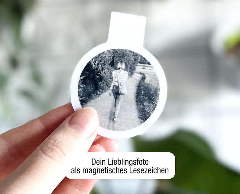 Ein magnetisches Lesezeichen mit Foto einer Person, die von hinten zu sehen ist, wird vor einem unscharfen Hintergrund in einer Hand gehalten. Darunter steht in einem weißen Feld: "Dein Lieblingsfoto als magnetisches Lesezeichen“.