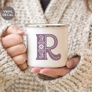 May include: White enamel mug with a purple vinyl decal of the letter "R" with floral embellishments. The decal is on the front of the mug.