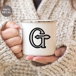Peut inclure: Mug en émail blanc avec un autocollant en vinyle noir et gris de la lettre "G" avec une flèche pointant vers la droite. L'autocollant est sur le devant de la tasse.