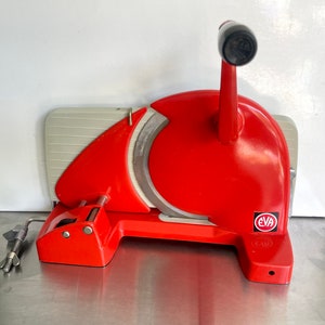 May include: A red and white manual meat slicer with a black handle. The slicer has a silver blade and a white plastic base. The brand name "EVA" is printed on the side of the slicer.