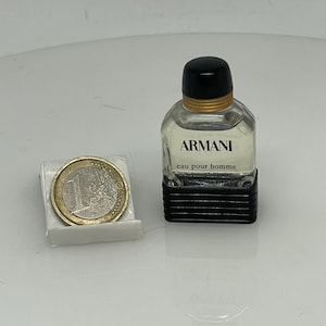 Puede incluir: Un peque&ntilde;o frasco de colonia Armani eau pour homme de vidrio transparente. El frasco tiene una tapa negra y una etiqueta negra con texto blanco. El frasco est&aacute; sobre una superficie blanca junto a una moneda de euro.