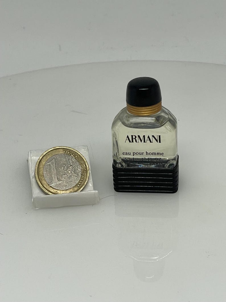 Puede incluir: Un peque&ntilde;o frasco de colonia Armani eau pour homme de vidrio transparente. El frasco tiene una tapa negra y una etiqueta negra con texto blanco. El frasco est&aacute; sobre una superficie blanca junto a una moneda de euro.