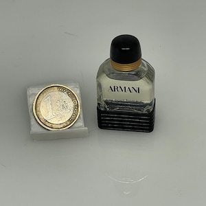 Puede incluir: Un peque&ntilde;o frasco de colonia Armani de vidrio transparente con tapa negra y banda dorada. El frasco est&aacute; sobre una superficie blanca junto a una moneda de euro.