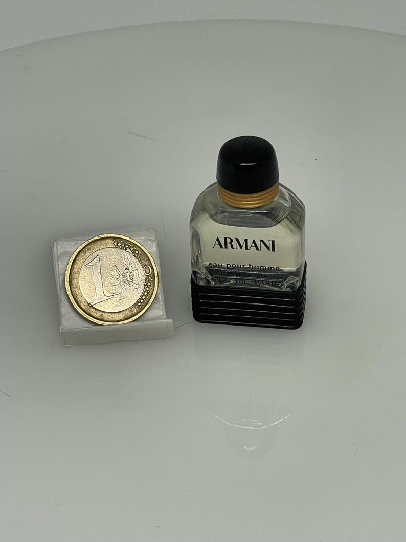 Puede incluir: Un peque&ntilde;o frasco de colonia Armani de vidrio transparente con tapa negra y banda dorada. El frasco est&aacute; sobre una superficie blanca junto a una moneda de euro.