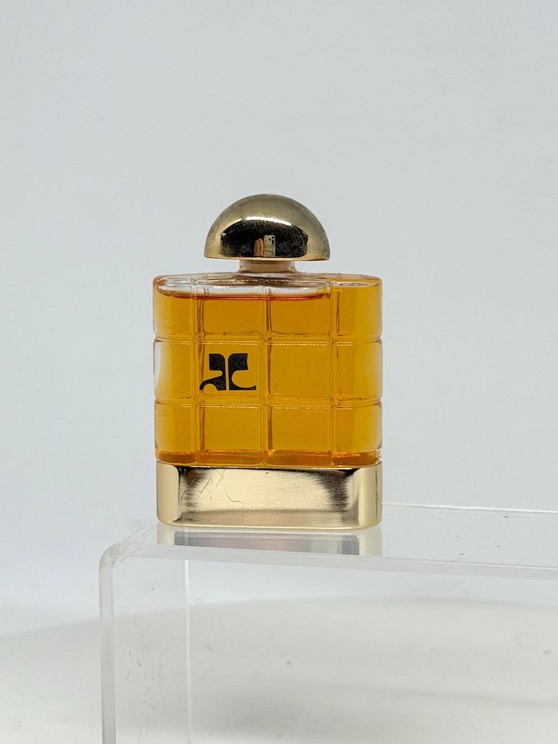 Peut inclure: Un petit flacon de parfum carr&eacute; avec un bouchon et une base dor&eacute;s. La bouteille est de couleur ambre chaud et pr&eacute;sente un motif quadrill&eacute; sur le devant. Un logo noir est visible sur le devant.