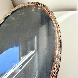 May include: Oval-shaped antique picture frame with a gold-colored ornate border. The frame has a dark interior and a glass cover. The frame's edge has a woven brown trim. The frame is likely vintage.