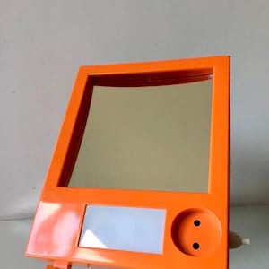 Op de afbeelding: Oranje kunststof make-upspiegel met een ingebouwd lampje en een stopcontact. De spiegel is schuin geplaatst.
