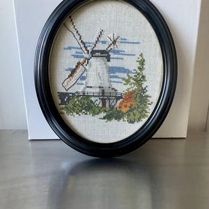 May include: A framed cross-stitch embroidery depicting a windmill with a blue sky and green trees. The frame is black and oval shaped.