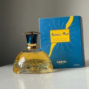 May include: A blue and yellow box with the text "Aimez-Moi Eau de Toilette Natural Spray" and the brand name "CARON PARIS" on the front. The box contains a clear glass bottle of perfume with a gold cap and the brand name "CARON PARIS" on the bottle. The bottle is 3.3 fl oz.