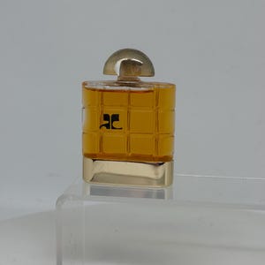 Peut inclure: Un petit flacon de parfum carr&eacute; avec une base dor&eacute;e et un bouchon transparent arrondi. Le flacon est rempli d'un liquide dor&eacute; et porte un logo noir sur le devant. Le flacon est pr&eacute;sent&eacute; sur une surface en acrylique transparente.