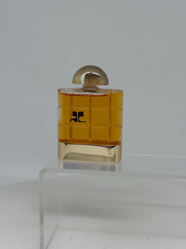 Peut inclure: Un petit flacon de parfum carr&eacute; avec une base dor&eacute;e et un bouchon transparent arrondi. Le flacon est rempli d'un liquide dor&eacute; et porte un logo noir sur le devant. Le flacon est pr&eacute;sent&eacute; sur une surface en acrylique transparente.