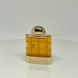 Peut inclure: Un petit flacon de parfum rectangulaire avec un bouchon et une base dor&eacute;s. Le corps du flacon pr&eacute;sente un motif quadrill&eacute; de couleur ambre chaud. Le logo "EC" est visible &agrave; l'avant. Le flacon se refl&egrave;te sur la surface en dessous.