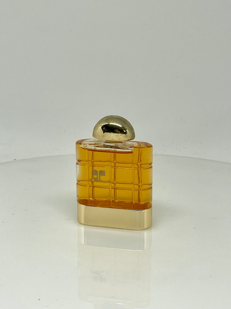Peut inclure: Un petit flacon de parfum rectangulaire avec un bouchon et une base dor&eacute;s. Le corps du flacon pr&eacute;sente un motif quadrill&eacute; de couleur ambre chaud. Le logo "EC" est visible &agrave; l'avant. Le flacon se refl&egrave;te sur la surface en dessous.