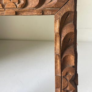 Op de afbeelding: Detailopname van een hoek van een houten frame met ingewikkelde gesneden bloemen- en bladerpatronen. Het frame is gemaakt van lichtbruin hout en heeft een natuurlijke, rustieke uitstraling. De gravures voegen een decoratief element toe.