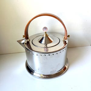 Könnte beinhalten: Ein polierter silberfarbener Metallkessel mit zylindrischem Körper und einer Reihe kleiner dekorativer Nieten am oberen Rand. Er verfügt über einen geschwungenen Holzgriff, einen konischen Deckel mit einem kleinen rosa Kugelknopf und einen kurzen Ausguss.