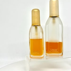 Könnte beinhalten: Zwei Glasparfümflaschen mit goldfarbenen Kappen und einer hellen, bernsteinfarbenen Flüssigkeit im Inneren. Die Flaschen sind mit "Clinique Wrappings" in goldfarbenen Buchstaben beschriftet. Die Flaschen stehen auf einem transparenten, abgewinkelten Regal vor weißem Hintergrund.