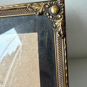 May include: Close-up of a vintage gold-toned picture frame. The frame has ornate corner details with a textured black background and a light-colored interior. The frame's edges have a dotted pattern.