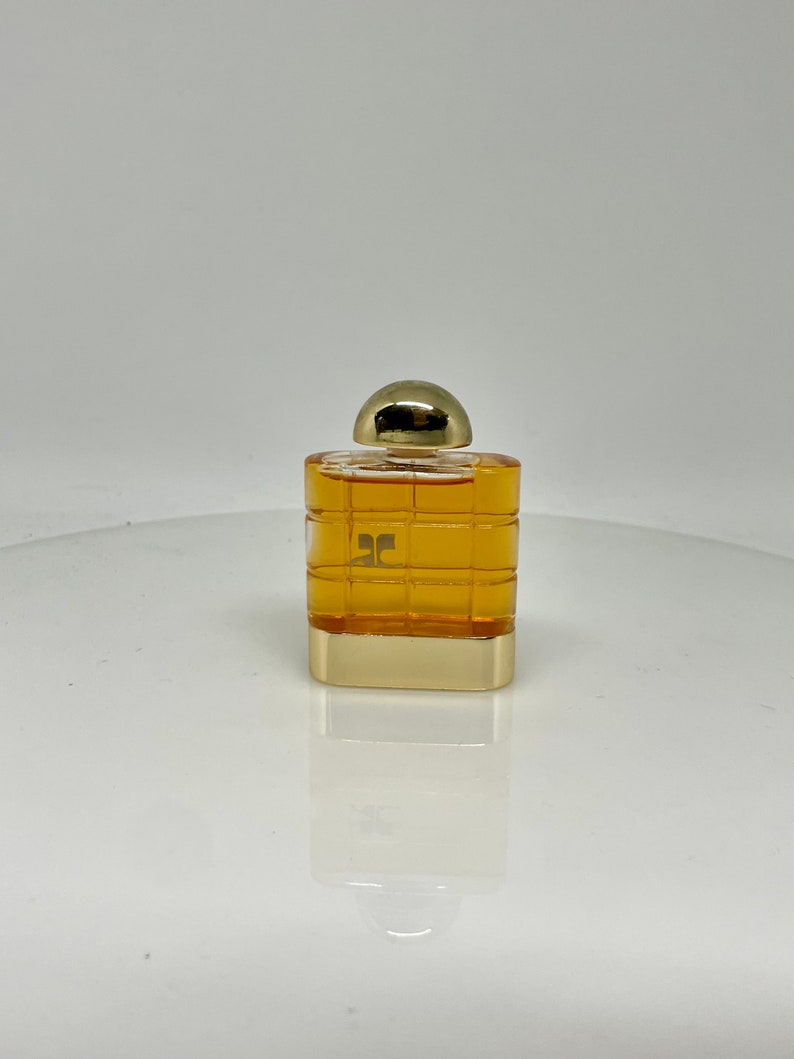 Peut inclure: Un petit flacon de parfum carr&eacute; avec un bouchon dor&eacute; et un logo dor&eacute;. La bouteille est remplie d'un liquide ambr&eacute; clair. La bouteille pr&eacute;sente un motif quadrill&eacute; sur les c&ocirc;t&eacute;s. La bouteille est pos&eacute;e sur une surface blanche.