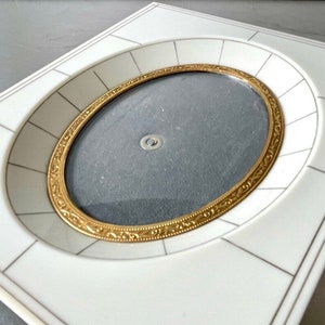 May include: Ivory-colored picture frame with an oval opening and a gold-colored ornate border. The frame has a rectangular shape with a decorative geometric pattern. The frame is designed to hold a photograph or artwork.