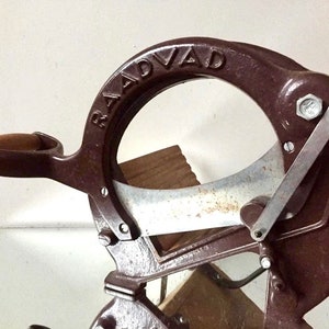 May include: A vintage brown cast iron RADVAD brand sausage slicer with a silver blade and a wooden handle.