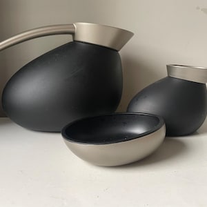 Puede incluir: Tres artículos de cocina de cerámica negra con detalles plateados. Una jarra grande con asa, una jarra más pequeña y un tazón.