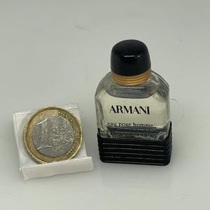 Puede incluir: Un peque&ntilde;o frasco de colonia Armani Eau Pour Homme de vidrio transparente. El frasco tiene una tapa negra y una banda dorada alrededor del cuello. El frasco est&aacute; sobre una superficie blanca junto a una moneda de euro.