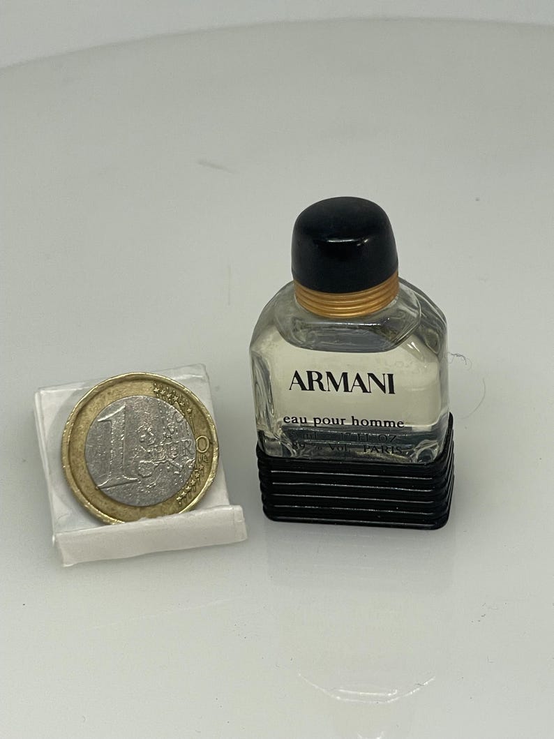 Puede incluir: Un peque&ntilde;o frasco de colonia Armani Eau Pour Homme de vidrio transparente. El frasco tiene una tapa negra y una banda dorada alrededor del cuello. El frasco est&aacute; sobre una superficie blanca junto a una moneda de euro.