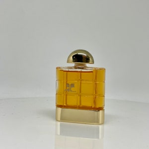 Peut inclure: Un flacon de parfum rectangulaire avec un bouchon et une base dor&eacute;s. Le flacon pr&eacute;sente un motif quadrill&eacute; et contient un liquide de couleur dor&eacute;e. Le logo "AC" est visible sur le devant du flacon.