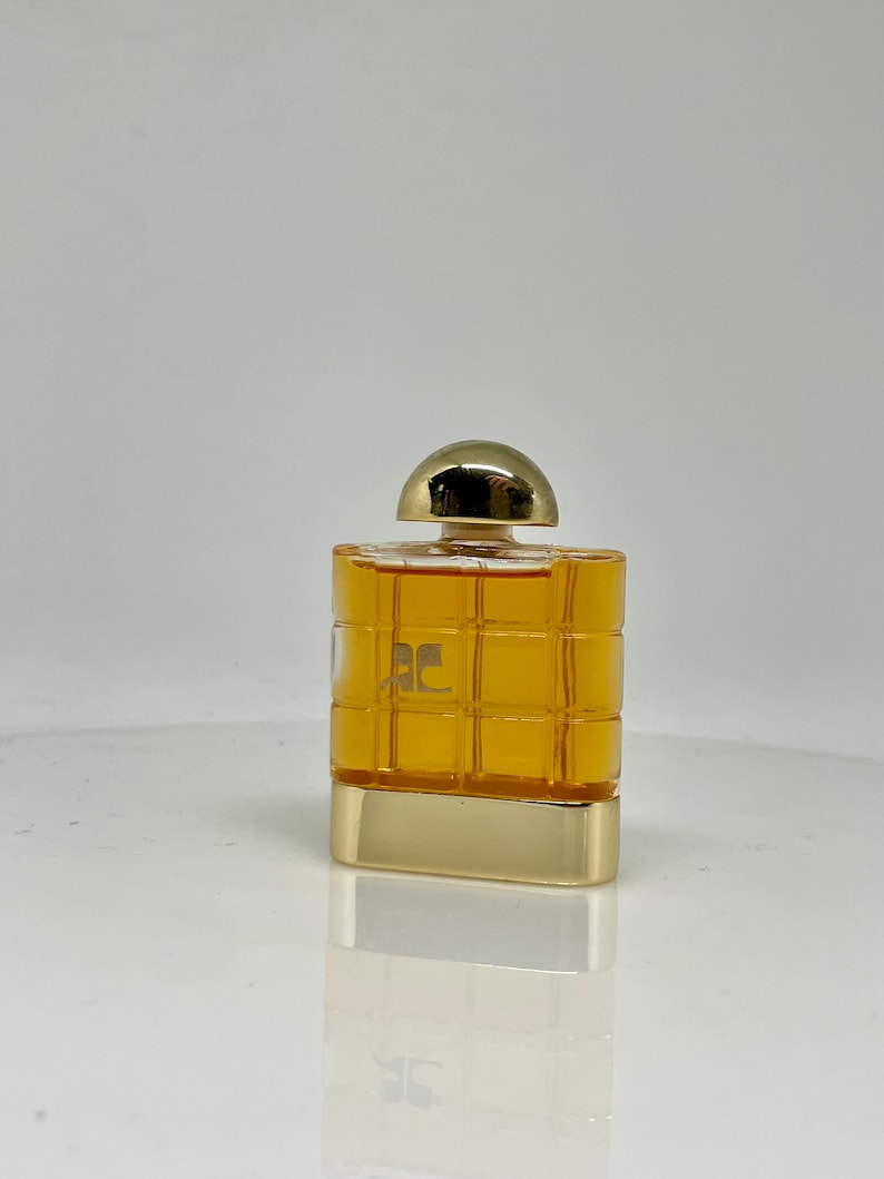 Peut inclure: Un flacon de parfum rectangulaire avec un bouchon et une base dor&eacute;s. Le flacon pr&eacute;sente un motif quadrill&eacute; et contient un liquide de couleur dor&eacute;e. Le logo "AC" est visible sur le devant du flacon.