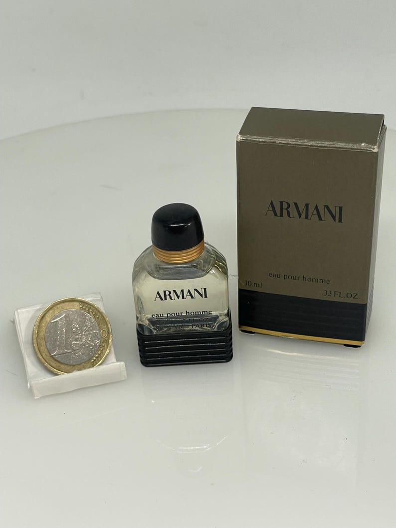 Puede incluir: Un peque&ntilde;o frasco de vidrio transparente de Armani Eau de Toilette para hombre. El frasco tiene una tapa negra y una banda dorada alrededor del cuello. El frasco est&aacute; sobre una superficie blanca junto a una moneda de euro. El frasco est&aacute; etiquetado con "Armani Eau pour Homme" y "10 ml". La caja es marr&oacute;n con letras doradas y dice "Armani Eau pour Homme" y "33 fl oz".