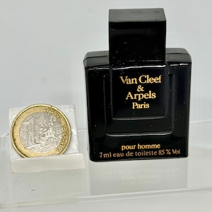 May include: A black rectangular perfume bottle with a square cap. The bottle has gold lettering that reads "Van Cleef & Arpels Paris pour homme". The bottle contains 7ml of eau de toilette, 85% Vol. A Euro coin is next to the bottle.