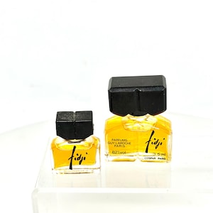 May include: Two miniature glass perfume bottles with amber liquid, black rectangular caps, and the text Fidji Parfums Guy Laroche Paris 82%vol 3.5 ml Cospar Paris printed on the larger bottle, while the smaller bottle features the text Fidji.